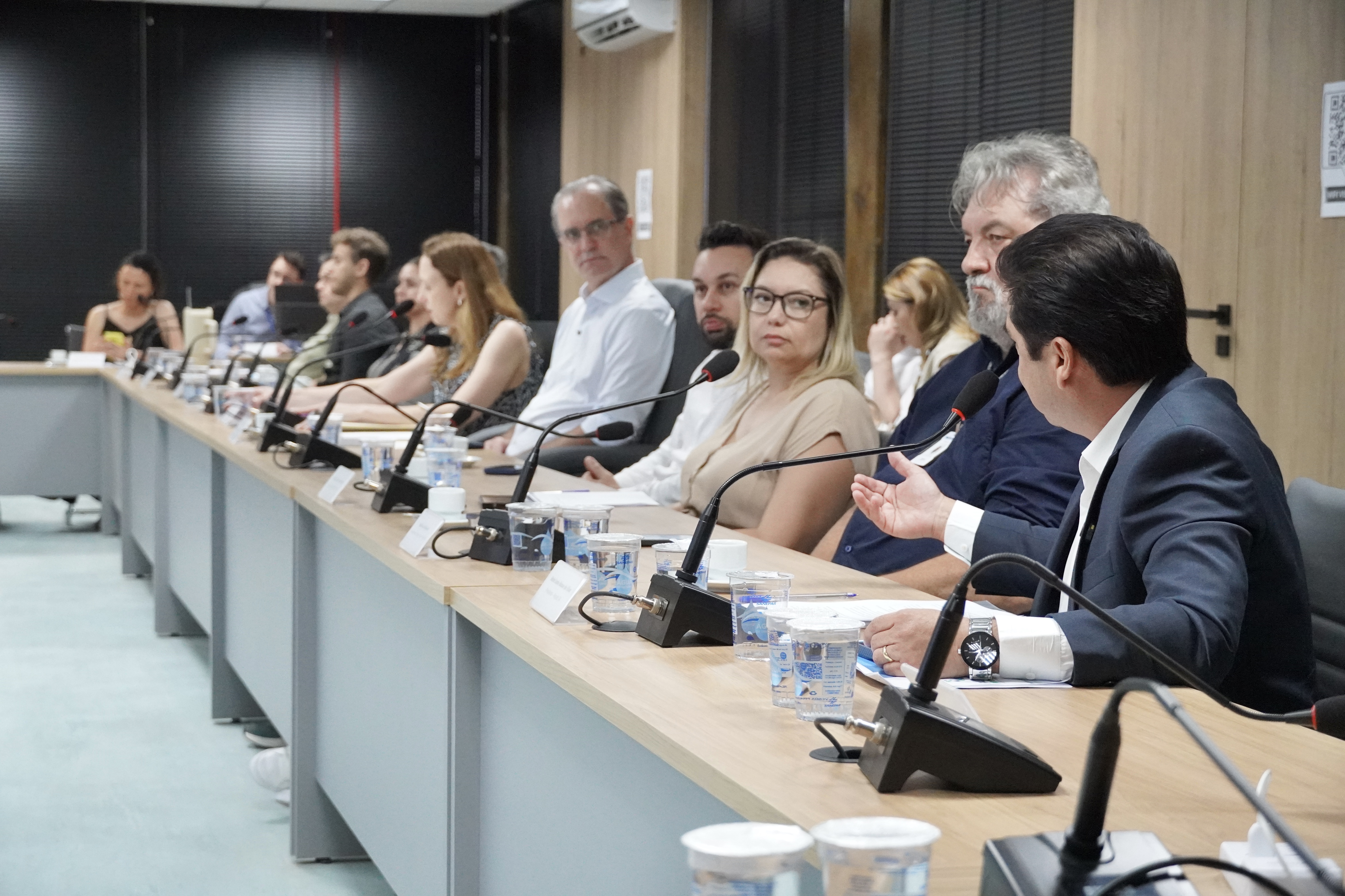 Longa mesa de reunião com várias pessoas sentadas em linha; em primeiro plano, um homem de terno gesticula ao falar ao microfone.