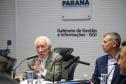 Fotografia foca no Vice-Governador Darci Piana, de terno bege e cabelos brancos, falando ao microfone e gesticulando com a mão direita. Sentado à sua esquerda, Luizão Goulart, secretário da Administração e da Previdência, de paletó azul, observa a fala. Ao fundo, a parede branca exibe o brasão do Governo do Paraná e a inscrição "Gabinete de Gestão e Informações - GGI". Em primeiro plano, elementos da mesa e participantes aparecem desfocados.