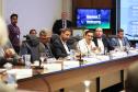 Fotografia de perfil lateral mostra uma fileira de homens sentados à mesa de reunião. Em destaque, ao centro, um homem de camisa branca gesticula com as mãos enquanto fala. Os demais participantes, vestidos formalmente, ouvem atentamente ou olham para baixo. Sobre a mesa de madeira, há placas de identificação, copos de água e microfones. Ao fundo, um monitor exibe o logotipo do Governo do Paraná e a inscrição "Gabinete de Gestão e Informações - GGI".