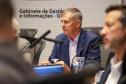 Fotografia foca em Luizão Goulart, secretário da Administração e da Previdência, sentado à mesa de reunião. Ele é um homem de pele clara e cabelos grisalhos curtos, vestindo paletó azul-marinho e camisa social clara. Ele olha para a esquerda da imagem com expressão atenta e segura uma caneta. À sua frente, microfones e copos d'água. Ao fundo, uma parede branca exibe o texto: 'Gabinete de Gestão e Informações - GGI'. Outras pessoas desfocadas aparecem nas laterais.