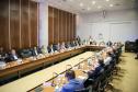 Fotografia em plano aberto mostra uma reunião formal em um auditório. Homens e mulheres estão sentados ao redor de uma grande mesa de madeira em formato de "U". Eles vestem trajes sociais e casuais finos. Sobre a mesa, há microfones, copos de água e papéis de anotação. Ao fundo, uma parede branca exibe o brasão do Governo do Estado do Paraná com a inscrição "Gabinete de Gestão e Informações - GGI". As paredes laterais são revestidas de madeira escura.