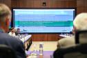 Fotografia em perspectiva central mostra um grande painel de LED ao fundo da sala de reuniões. O slide exibido tem o título "Próximos Passos" e detalha tópicos sobre os Conselhos Regionais e apoio na divulgação. À frente do telão, participantes sentados à mesa de madeira são vistos lateralmente. No primeiro plano, as costas e nucas de dois participantes estão desfocadas, emoldurando a imagem.