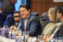 Fotografia em plano médio mostra um homem de cabelos escuros e paletó azul sorrindo e falando durante a reunião. Ele está sentado à mesa, rodeado por outros participantes que aparecem parcialmente. À sua frente, há microfones e copos de água sobre a mesa de madeira. O fundo apresenta painéis de madeira escura.