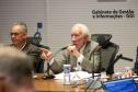 Fotografia foca em Darci Piana, Vice-Governador, de terno bege e cabelos brancos, falando ao microfone enquanto aponta com o dedo indicador da mão direita. Ele está sentado à mesa de reunião, olhando para a esquerda da imagem. À sua esquerda, sentado ao seu lado, está Domingos Trevisan Filho, diretor-geral da SEPL, de óculos e terno marrom, ouvindo atentamente. Sobre a mesa, há copos d'água e microfones. Ao fundo, uma parede branca exibe o texto: 'Gabinete de Gestão e Informações - GGI'.
