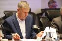 Fotografia em plano médio foca em Luizão Goulart, secretário da Administração e da Previdência, sentado à mesa de reunião. Ele está com a cabeça levemente abaixada, focado na leitura de documentos. Ele veste paletó azul-marinho sobre camisa social clara. Segura uma caneta na mão direita sobre um papel e manuseia outras folhas com a mão esquerda. À sua frente, copos d'água e uma xícara de café. Outros participantes desfocados estão ao seu redor.