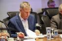 Fotografia em plano médio foca em Luizão Goulart, secretário da Administração e da Previdência, sentado à mesa de reunião. Ele está com os cabelos grisalhos abaixados, lendo atentamente documentos impressos sobre a mesa. Veste paletó azul-marinho e camisa social clara. Segura uma caneta na mão direita e folheia o papel com a esquerda. À sua frente, copos d'água de vidro e plástico, e uma xícara de café vazia. Ao fundo, desfocadas, aparecem outras pessoas sentadas à mesa.