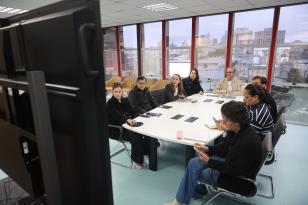 Pessoas sentadas ao redor de uma grande mesa branca em uma sala de reuniões envidraçada, participando de uma videoconferência exibida em um grande monitor em primeiro plano.