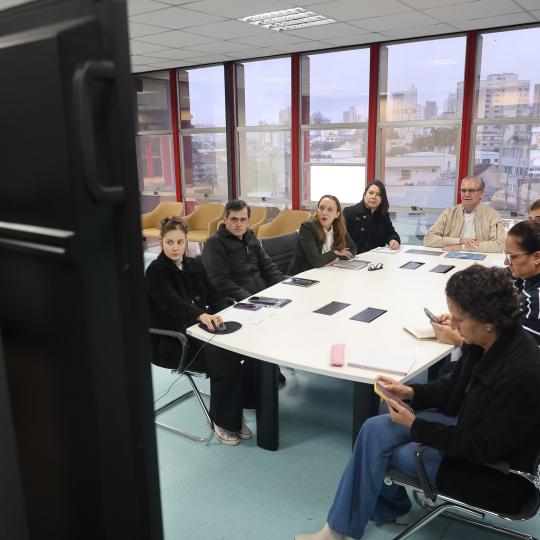 Pessoas sentadas ao redor de uma grande mesa branca em uma sala de reuniões envidraçada, participando de uma videoconferência exibida em um grande monitor em primeiro plano.