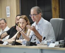 Close de um homem de camisa branca e óculos falando ao microfone em uma mesa de conferência; outras quatro pessoas aparecem ao fundo.