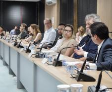 Longa mesa de reunião com várias pessoas sentadas em linha; em primeiro plano, um homem de terno gesticula ao falar ao microfone.