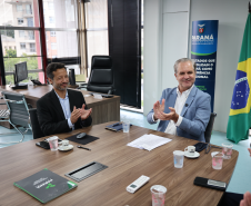 Dois homens sentados à mesa aplaudem; ao fundo, a bandeira do Brasil e um banner do Governo do Paraná.