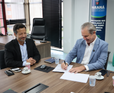 Homem de terno azul assina um documento à mesa enquanto outro homem ao lado observa sorrindo.