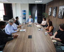 Visão ampla da mesa de reunião com nove pessoas sentadas; o homem de terno azul está na cabeceira, entre bandeiras e um banner.