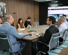 Reunião vista de perfil; várias pessoas sentadas à mesa e uma TV ao fundo exibindo um portal de notícias.