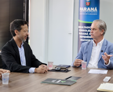 Dois homens conversam sentados; o da direita, de terno azul, gesticula. Ao fundo, um banner institucional e a bandeira nacional.