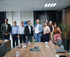 Foto de grupo em escritório formal. No centro, um homem de terno azul segura um documento oficial à frente de uma mesa de reunião. Ao fundo, aparecem as bandeiras do Brasil e do Paraná.