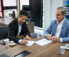 Homem de terno preto assina um documento enquanto o homem de terno azul ao lado acompanha o ato.