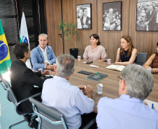 Seis pessoas ao redor da mesa em reunião; o homem de terno azul à esquerda lidera a conversa sob fotos institucionais.