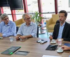 Três homens sentados; o da direita gesticula durante a fala. Ao fundo, janelas amplas com persianas.
