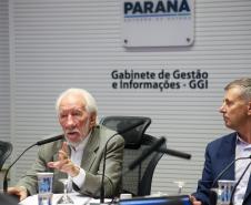Fotografia foca no Vice-Governador Darci Piana, de terno bege e cabelos brancos, falando ao microfone e gesticulando com a mão direita. Sentado à sua esquerda, Luizão Goulart, secretário da Administração e da Previdência, de paletó azul, observa a fala. Ao fundo, a parede branca exibe o brasão do Governo do Paraná e a inscrição "Gabinete de Gestão e Informações - GGI". Em primeiro plano, elementos da mesa e participantes aparecem desfocados.