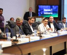 Fotografia de perfil lateral mostra uma fileira de homens sentados à mesa de reunião. Em destaque, ao centro, um homem de camisa branca gesticula com as mãos enquanto fala. Os demais participantes, vestidos formalmente, ouvem atentamente ou olham para baixo. Sobre a mesa de madeira, há placas de identificação, copos de água e microfones. Ao fundo, um monitor exibe o logotipo do Governo do Paraná e a inscrição "Gabinete de Gestão e Informações - GGI".