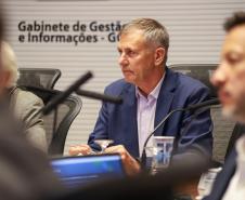 Fotografia foca em Luizão Goulart, secretário da Administração e da Previdência, sentado à mesa de reunião. Ele é um homem de pele clara e cabelos grisalhos curtos, vestindo paletó azul-marinho e camisa social clara. Ele olha para a esquerda da imagem com expressão atenta e segura uma caneta. À sua frente, microfones e copos d'água. Ao fundo, uma parede branca exibe o texto: 'Gabinete de Gestão e Informações - GGI'. Outras pessoas desfocadas aparecem nas laterais.