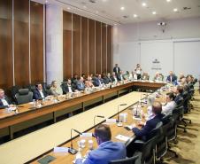 Fotografia em plano aberto mostra uma reunião formal em um auditório. Homens e mulheres estão sentados ao redor de uma grande mesa de madeira em formato de "U". Eles vestem trajes sociais e casuais finos. Sobre a mesa, há microfones, copos de água e papéis de anotação. Ao fundo, uma parede branca exibe o brasão do Governo do Estado do Paraná com a inscrição "Gabinete de Gestão e Informações - GGI". As paredes laterais são revestidas de madeira escura.