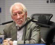 Fotografia em plano médio do vice-governador, um homem branco, de cabelos e barba curtos e brancos. Ele veste paletó cinza xadrez sobre camisa branca e está sentado em uma cadeira de escritório preta. À sua frente, sobre a mesa, há um microfone e copos d'água. Ele olha para a direita com expressão atenta e séria.