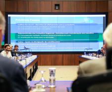 Fotografia em perspectiva central mostra um grande painel de LED ao fundo da sala de reuniões. O slide exibido tem o título "Próximos Passos" e detalha tópicos sobre os Conselhos Regionais e apoio na divulgação. À frente do telão, participantes sentados à mesa de madeira são vistos lateralmente. No primeiro plano, as costas e nucas de dois participantes estão desfocadas, emoldurando a imagem.