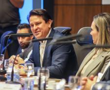 Fotografia em plano médio mostra um homem de cabelos escuros e paletó azul sorrindo e falando durante a reunião. Ele está sentado à mesa, rodeado por outros participantes que aparecem parcialmente. À sua frente, há microfones e copos de água sobre a mesa de madeira. O fundo apresenta painéis de madeira escura.