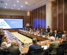 Reunião formal do Conselho Gestor Estadual em auditório amplo. Mais de vinte pessoas, em trajes sociais e casuais finos, estão sentadas ao redor de uma grande mesa de madeira em formato de U. Ao fundo, um grande painel de LED exibe o slide da "4ª Reunião do Conselho Gestor Estadual", com o logotipo "Paraná Produtivo".