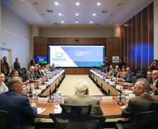 Reunião presencial do Conselho Gestor Estadual em auditório com grande mesa em formato de U. Mais de 20 participantes, vestindo trajes sociais e casuais finos, estão sentados ao redor da mesa. Ao fundo, um painel de LED exibe o slide da "4ª Reunião do Conselho Gestor Estadual", com o logotipo "Paraná Produtivo".
