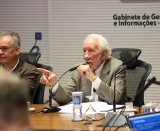 Fotografia foca em Darci Piana, Vice-Governador, de terno bege e cabelos brancos, falando ao microfone enquanto aponta com o dedo indicador da mão direita. Ele está sentado à mesa de reunião, olhando para a esquerda da imagem. À sua esquerda, sentado ao seu lado, está Domingos Trevisan Filho, diretor-geral da SEPL, de óculos e terno marrom, ouvindo atentamente. Sobre a mesa, há copos d'água e microfones. Ao fundo, uma parede branca exibe o texto: 'Gabinete de Gestão e Informações - GGI'.