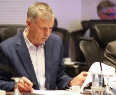 Fotografia em plano médio foca em Luizão Goulart, secretário da Administração e da Previdência, sentado à mesa de reunião. Ele está com a cabeça levemente abaixada, focado na leitura de documentos. Ele veste paletó azul-marinho sobre camisa social clara. Segura uma caneta na mão direita sobre um papel e manuseia outras folhas com a mão esquerda. À sua frente, copos d'água e uma xícara de café. Outros participantes desfocados estão ao seu redor.