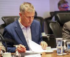 Fotografia em plano médio foca em Luizão Goulart, secretário da Administração e da Previdência, sentado à mesa de reunião. Ele está com os cabelos grisalhos abaixados, lendo atentamente documentos impressos sobre a mesa. Veste paletó azul-marinho e camisa social clara. Segura uma caneta na mão direita e folheia o papel com a esquerda. À sua frente, copos d'água de vidro e plástico, e uma xícara de café vazia. Ao fundo, desfocadas, aparecem outras pessoas sentadas à mesa.