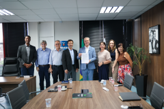 Foto de grupo em escritório formal. No centro, um homem de terno azul segura um documento oficial à frente de uma mesa de reunião. Ao fundo, aparecem as bandeiras do Brasil e do Paraná.