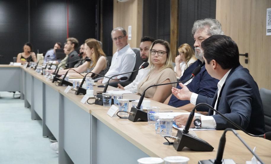 Longa mesa de reunião com várias pessoas sentadas em linha; em primeiro plano, um homem de terno gesticula ao falar ao microfone.