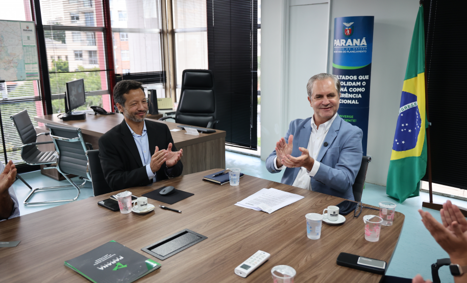 Dois homens sentados à mesa aplaudem; ao fundo, a bandeira do Brasil e um banner do Governo do Paraná.