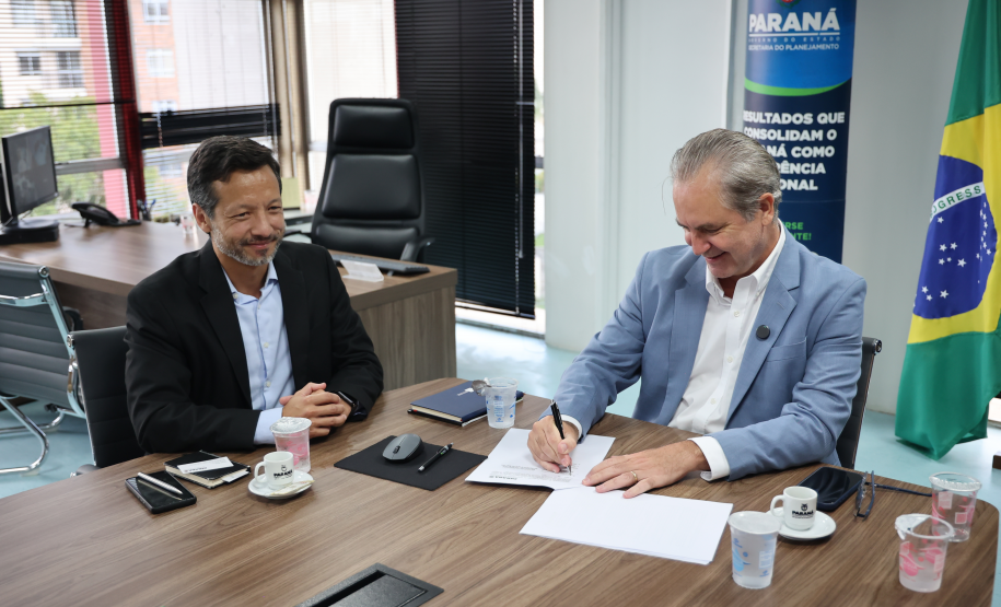 Homem de terno azul assina um documento à mesa enquanto outro homem ao lado observa sorrindo.