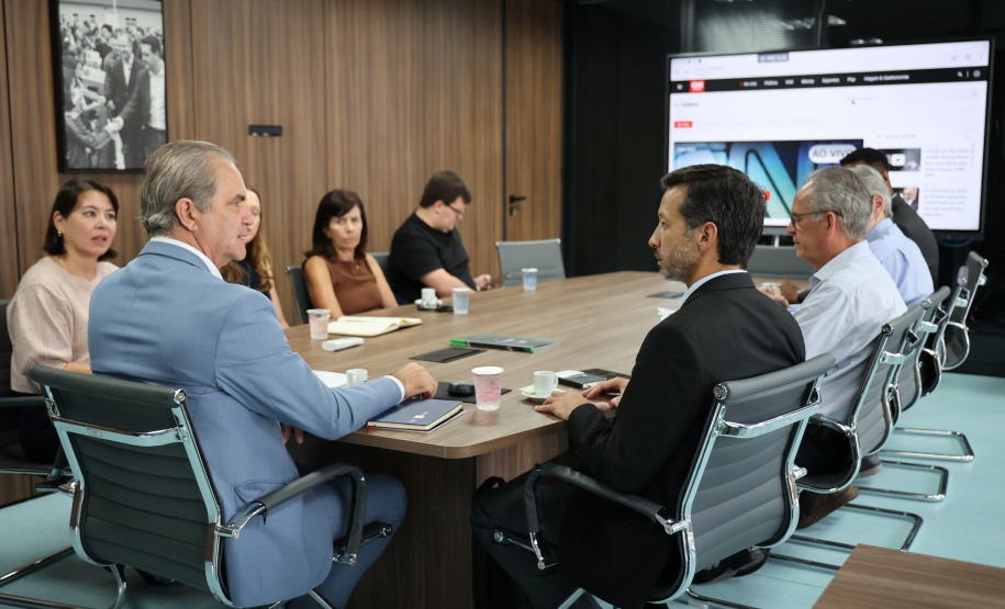 Reunião vista de perfil; várias pessoas sentadas à mesa e uma TV ao fundo exibindo um portal de notícias.