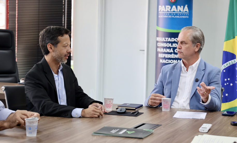 Dois homens conversam sentados; o da direita, de terno azul, gesticula. Ao fundo, um banner institucional e a bandeira nacional.