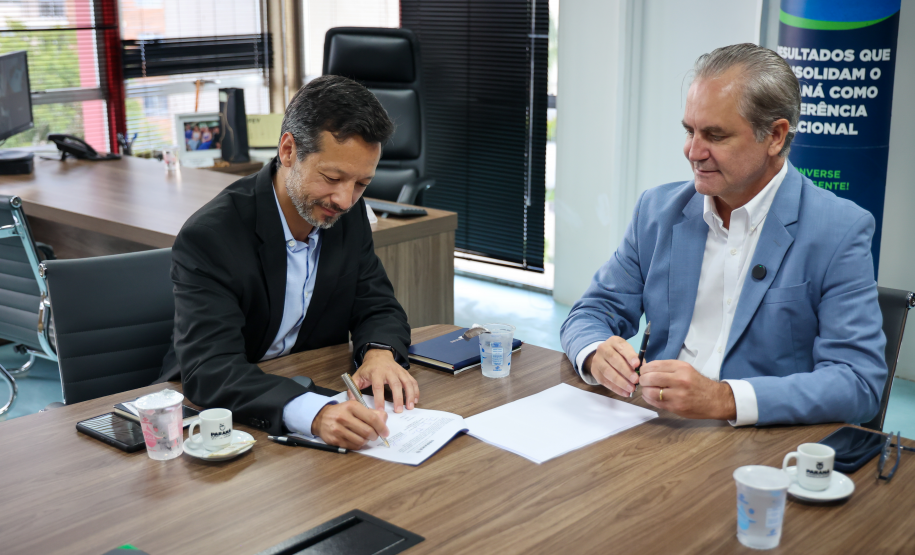 Homem de terno preto assina um documento enquanto o homem de terno azul ao lado acompanha o ato.