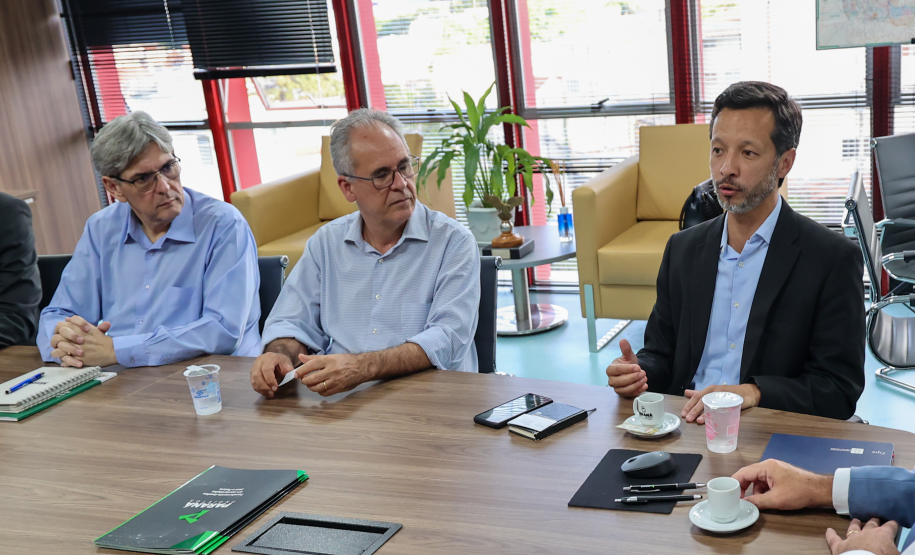 Três homens sentados; o da direita gesticula durante a fala. Ao fundo, janelas amplas com persianas.