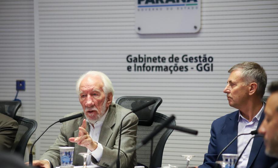 Fotografia foca no Vice-Governador Darci Piana, de terno bege e cabelos brancos, falando ao microfone e gesticulando com a mão direita. Sentado à sua esquerda, Luizão Goulart, secretário da Administração e da Previdência, de paletó azul, observa a fala. Ao fundo, a parede branca exibe o brasão do Governo do Paraná e a inscrição "Gabinete de Gestão e Informações - GGI". Em primeiro plano, elementos da mesa e participantes aparecem desfocados.