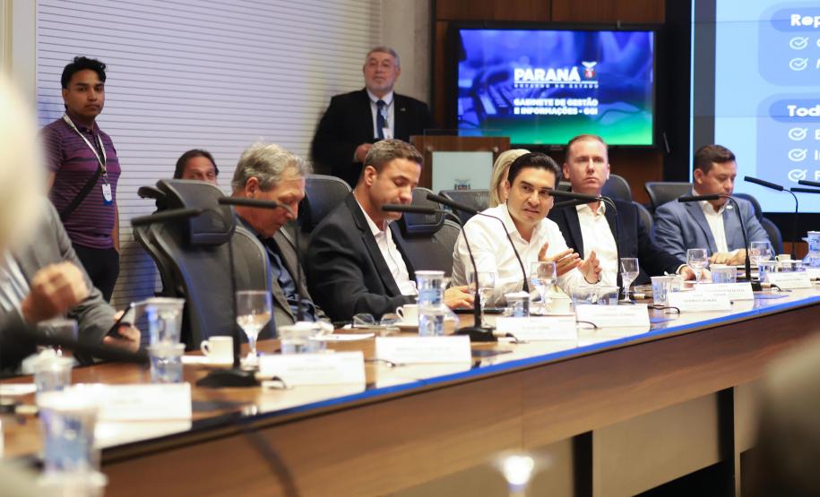 Fotografia de perfil lateral mostra uma fileira de homens sentados à mesa de reunião. Em destaque, ao centro, um homem de camisa branca gesticula com as mãos enquanto fala. Os demais participantes, vestidos formalmente, ouvem atentamente ou olham para baixo. Sobre a mesa de madeira, há placas de identificação, copos de água e microfones. Ao fundo, um monitor exibe o logotipo do Governo do Paraná e a inscrição "Gabinete de Gestão e Informações - GGI".