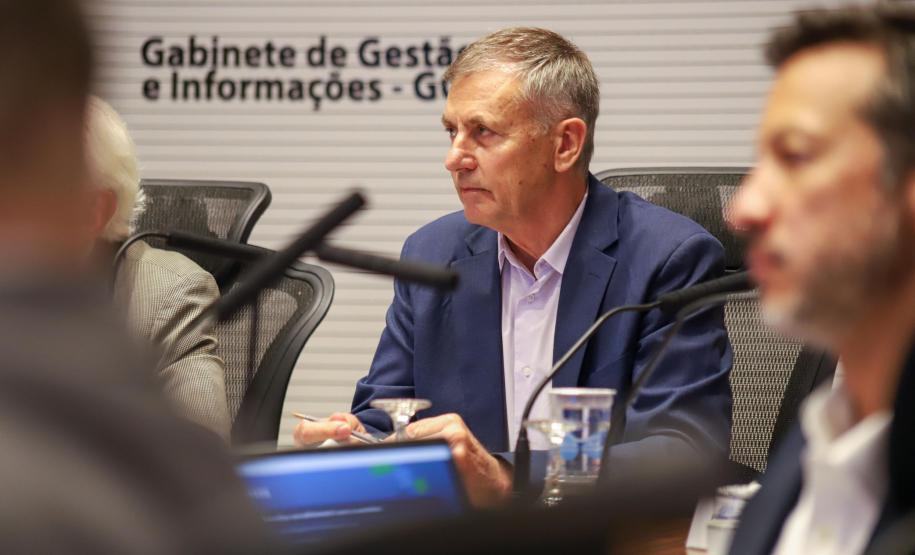 Fotografia foca em Luizão Goulart, secretário da Administração e da Previdência, sentado à mesa de reunião. Ele é um homem de pele clara e cabelos grisalhos curtos, vestindo paletó azul-marinho e camisa social clara. Ele olha para a esquerda da imagem com expressão atenta e segura uma caneta. À sua frente, microfones e copos d'água. Ao fundo, uma parede branca exibe o texto: 'Gabinete de Gestão e Informações - GGI'. Outras pessoas desfocadas aparecem nas laterais.