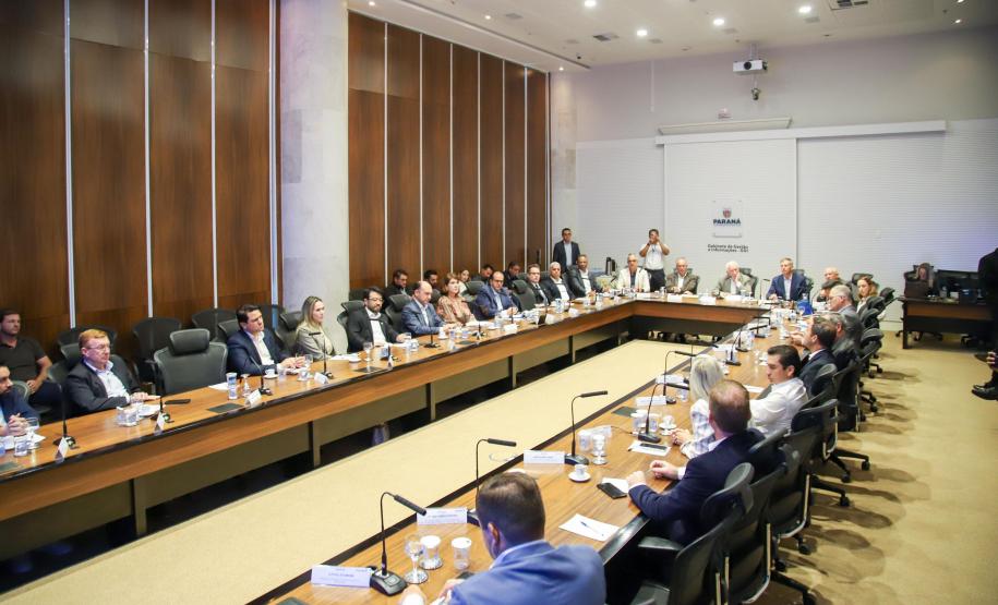 Fotografia em plano aberto mostra uma reunião formal em um auditório. Homens e mulheres estão sentados ao redor de uma grande mesa de madeira em formato de "U". Eles vestem trajes sociais e casuais finos. Sobre a mesa, há microfones, copos de água e papéis de anotação. Ao fundo, uma parede branca exibe o brasão do Governo do Estado do Paraná com a inscrição "Gabinete de Gestão e Informações - GGI". As paredes laterais são revestidas de madeira escura.