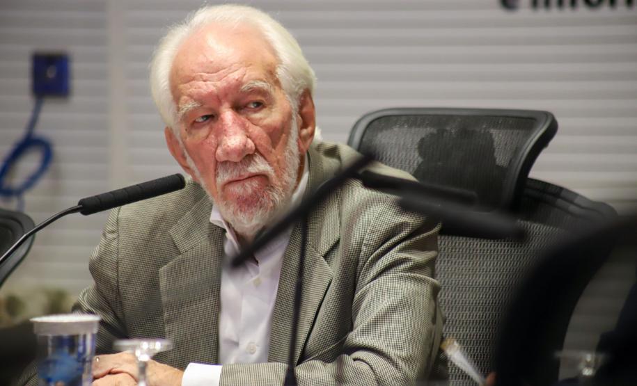 Fotografia em plano médio do vice-governador, um homem branco, de cabelos e barba curtos e brancos. Ele veste paletó cinza xadrez sobre camisa branca e está sentado em uma cadeira de escritório preta. À sua frente, sobre a mesa, há um microfone e copos d'água. Ele olha para a direita com expressão atenta e séria.