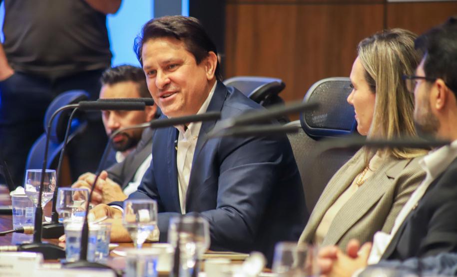 Fotografia em plano médio mostra um homem de cabelos escuros e paletó azul sorrindo e falando durante a reunião. Ele está sentado à mesa, rodeado por outros participantes que aparecem parcialmente. À sua frente, há microfones e copos de água sobre a mesa de madeira. O fundo apresenta painéis de madeira escura.