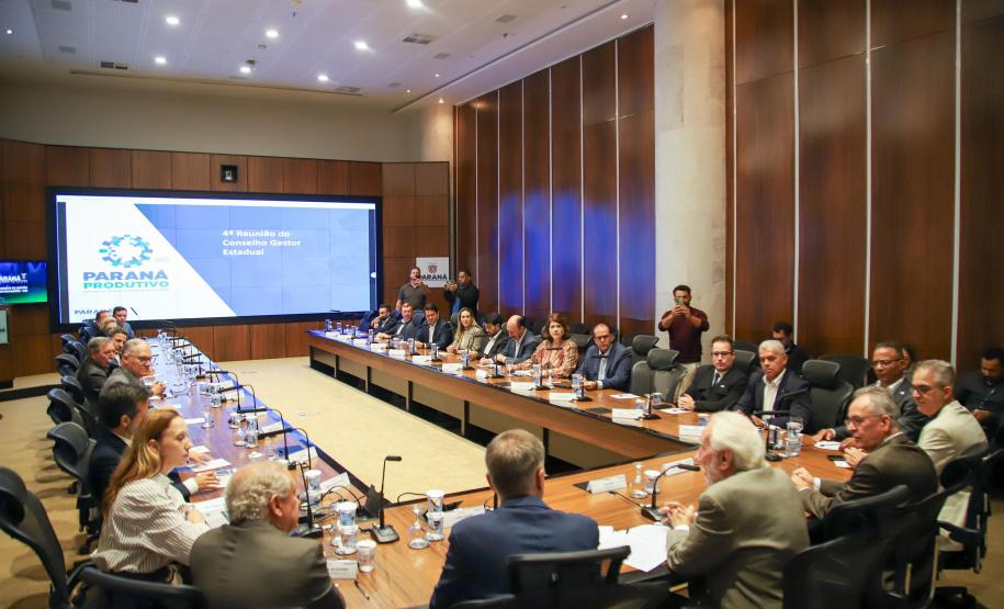 Reunião formal do Conselho Gestor Estadual em auditório amplo. Mais de vinte pessoas, em trajes sociais e casuais finos, estão sentadas ao redor de uma grande mesa de madeira em formato de U. Ao fundo, um grande painel de LED exibe o slide da "4ª Reunião do Conselho Gestor Estadual", com o logotipo "Paraná Produtivo".